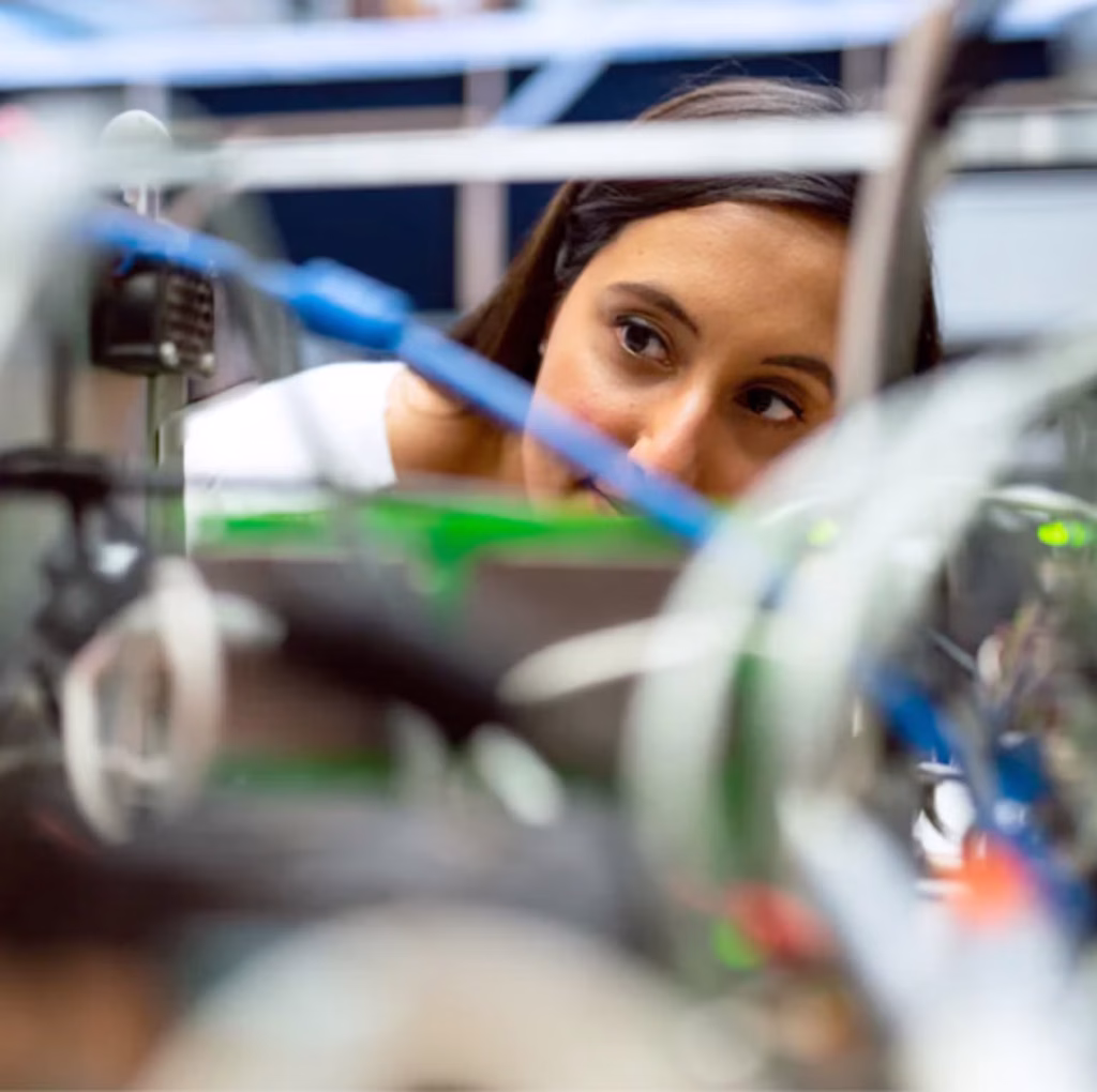 A woman intently observing a computer in a factory, with industrial equipment visible in the background.
