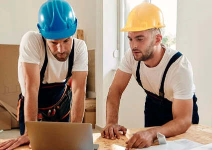 Two men wearing hard hats are engaged in a discussion while working on a laptop at a construction site.