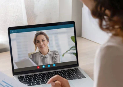 A woman views a video of another woman on her laptop screen while seated at a desk.
